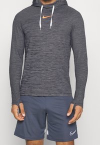 Nike Langarm-Hoodie, dunkelgrau mit rotem Logo, aus strukturiertem Stoff, weißen Kordeln und eng anliegenden Bündchen. Getragen mit blauen Shorts.