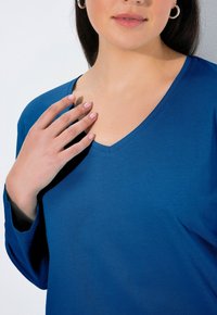 Niebieski top z dekoltem w serek wykonany z gładkiej, miękkiej tkaniny. Długie rękawy o luźnym kroju, z czystym wykończeniem przy dekolcie i prostym designem.