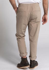 Beige, luchtige broeken met een elastische tailleband, twee achterzakken en opgerolde boorden. Draagt een paar donkere veterschoenen.