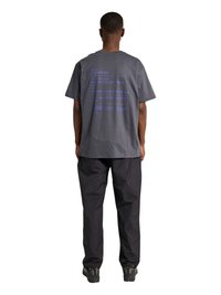 Man staat met zijn rug naar de camera, draagt een grafisch charcoal grijs t-shirt, zwarte broek en zwarte sneakers op een witte achtergrond.