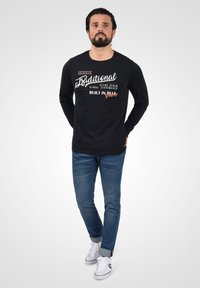 Sort langærmet skjorte med hvid og orange grafisk tekst på forsiden, parret med blå denim jeans og hvide sneakers. Model står med hænderne i lommerne.