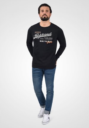 Camisa negra de manga larga con texto gráfico blanco y naranja en la parte frontal, combinada con jeans de mezclilla azul y zapatillas blancas. Modelo de pie con las manos en los bolsillos.