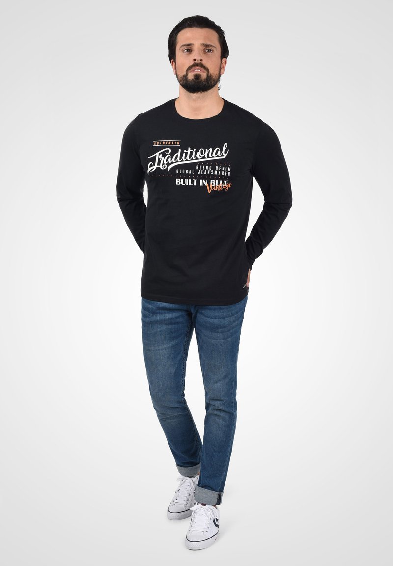 Sort langærmet skjorte med hvid og orange grafisk tekst på forsiden, parret med blå denim jeans og hvide sneakers. Model står med hænderne i lommerne.