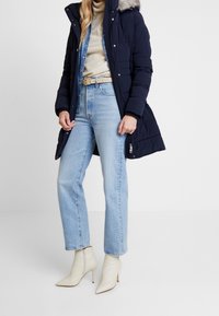 Giacca puffer blu navy con cappuccio in finto pelo, abbinata a jeans a vita alta azzurri chiari e stivaletti bianchi con punta. Texture morbide, design strutturato.