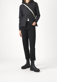 Mujer vistiendo una chaqueta negra ligera acolchada, pantalones negros cortos, botas gruesas negras y una correa de bolso cruzado blanca visible.