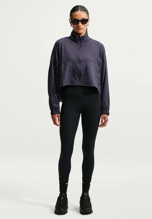 Vrouw met zwarte zonnebril, een donkerpaarse cropped jas met ritssluiting, zwarte legging, zwarte sokken en zwarte schoenen, staand tegen een effen achtergrond.