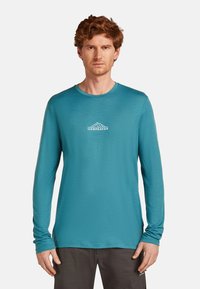 Langärmliges sportliches Shirt in Türkis, mit runder Ausschnitt und einem kleinen weißen Berg-Logo auf der Brust. Der Stoff hat eine glatte Textur.