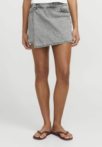 Gonna avvolgente in denim grigia, con orlo sfilacciato e passanti per cintura. Abbinata a ciabatte viola, che mettono in mostra le gambe scoperte.