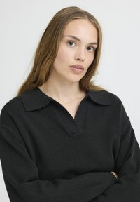 Juodas megztinis su V formos iškirpte ir apykakle, turintis laisvą siluetą ir ilgomis rankovėmis, pagamintas iš švelnaus tekstūrinio audinio.