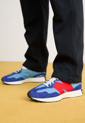 Blå ruskind og meshesneakers med røde og lyseblå accenter, med en hvid sål og snørebånd, set fra siden.