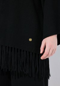 Schwarzer Poncho mit Fransen und einem goldenen Metalletikett. Weicher, strukturierter Stoff fällt elegant. Die Fransen verleihen Bewegung und visuelles Interesse.