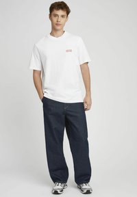 T-shirt en coton blanc avec le texte rouge "DISPONIBLE POUR UN RENDEZ-VOUS", associé à un pantalon en denim foncé ample et des baskets grises.