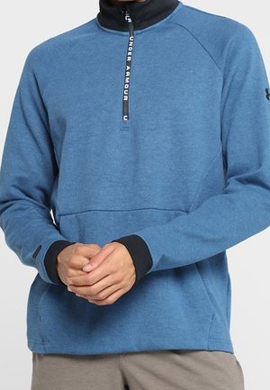 Mand iført blå Under Armour kvart-lynlås pullover med sort krave og manchetter, hænder foldet ved taljen, beige bukser synlige.