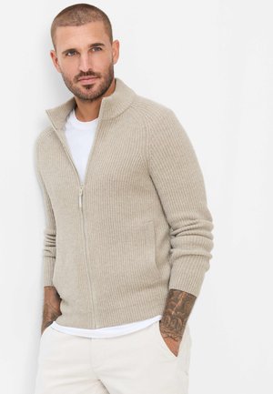 Uomo con capelli corti e avambracci tatuati che indossa un maglione beige con zip sopra una camicia bianca e pantaloni chiari, in piedi con le mani in tasca.