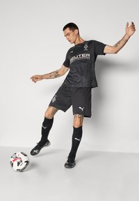 Svart fotbollsdräkt med en strukturerad design och vita logodetaljer. Spelare i rörelse bredvid en mönstrad fotboll på en ljus bakgrund.