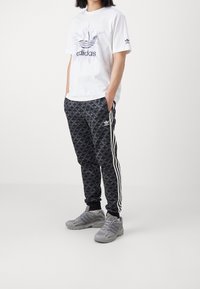 Bílá tričko s grafickým logem, kombinované s černými vzorovanými tepláky s bílými pruhy a šedými sportovními botami.