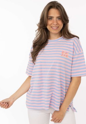 Frau mit langen braunen Haaren, die ein rosa-blau gestreiftes T-Shirt mit dem auf der Brust gestickten Text "BONJOUR" trägt, lächelnd, weißer Hintergrund.