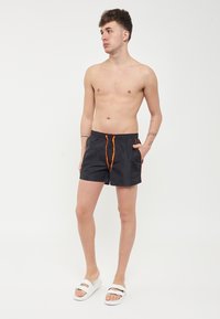 Costume da nuoto neri realizzati in materiale leggero, con cordoncino arancione e dettagli laterali. Indossati con sandali bianchi.