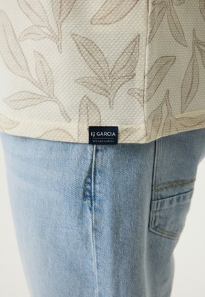 Beige gestructureerde stof met bladmotief en "Garcia" label, gecombineerd met lichtblauwe denim jeans met zijzakstiksel.