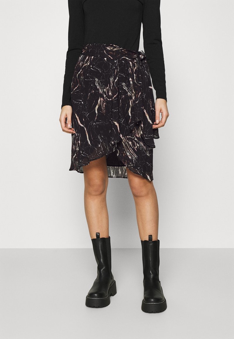 Femme portant un haut noir à manches longues, une jupe portefeuille à motif abstrait noir et blanc, et des bottes noires épaisses mi-mollet, debout sur un sol gris.