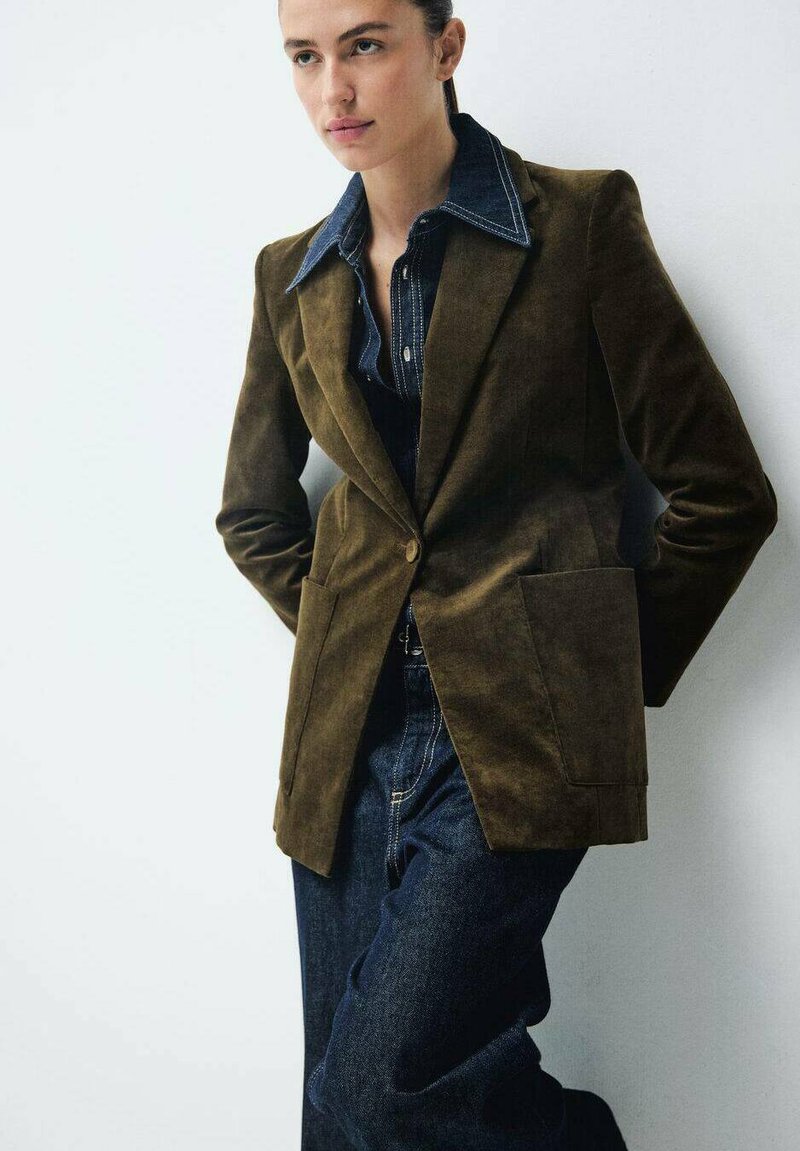 Blazer in velluto verde oliva con revers, due tasche frontali, abbinato a una camicia in denim scuro e jeans blu. Texture liscia, design strutturato.