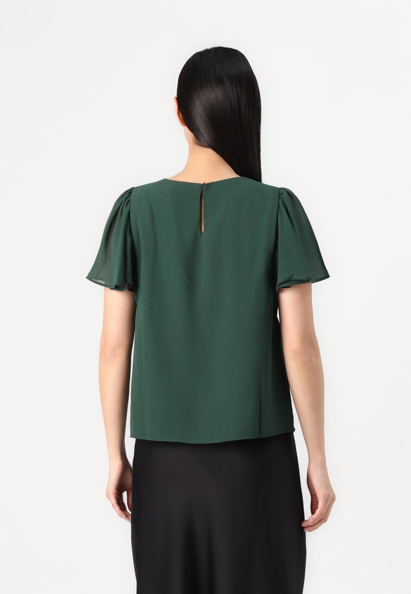 Blouse verte à manches courtes avec un col rond, dotée de manches en flutter douces et transparentes ainsi que d'un détail en forme de trou de serrure à l'arrière. Texture lisse.