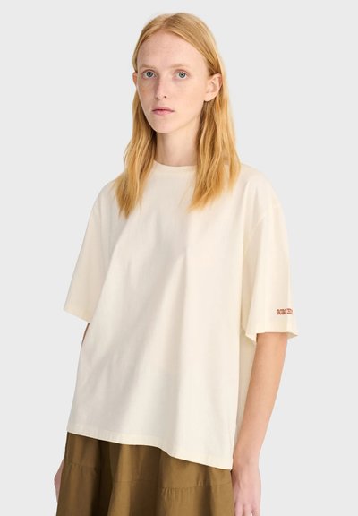 Jeune femme aux cheveux raides roux clair porte un t-shirt ample couleur crème et une jupe marron sur un fond clair uni.