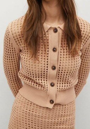 Femme portant un cardigan beige en tricot avec de grands trous et des boutons marron, assorti à une jupe taille haute.