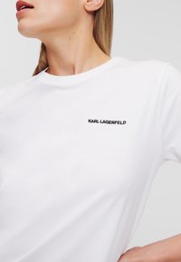 Bílá bavlněná tričko s kulatým výstřihem a krátkými rukávy, s malým černým logem "KARL LAGERFELD" na levé straně.