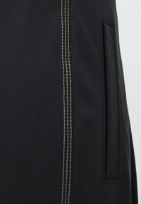 Zwart kledingstuk met een gestructureerd oppervlak, contrasterende lichtbeige stiksels en een zijzak ontworpen met een subtiele naad.