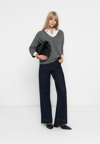 Pull en maille gris à col en V, chemise blanche à col en dessous, associé à un jean en denim foncé à jambes larges et des chaussures plates noires. Le mannequin tient un sac noir.