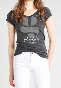 Šedé tričko s krátkým rukávem a zaobleným výstřihem, s tečkovaným designem písmene 'S' a tučným bílým nápisem "RAW". Nosené s bílými džíny.