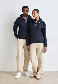 Mørkeblå pulloverjakker med hætte, der har en frontlomme og lynlås, parret med beige buksestil i vidde og hvide sneakers.
