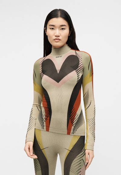 Γυναίκα με μακριά σκούρα μαλλιά που φοράει εφαρμοστή, εμπριμέ μακρυμάνικη μπλούζα και ταιριαστό παντελόνι σε μπεζ, μαύρους, κόκκινους, κίτρινους και ροζ τόνους.