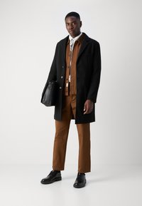 Black wool coat, brown button-up shirt, brown trousers, black leather shoes, holding a black bag. Clean design, tailored fit.