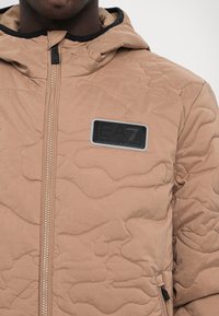 Quiltet beige jakke med hætte, der har et camouflage mønster, en sort mærke, og en synlig lynlås i midten.