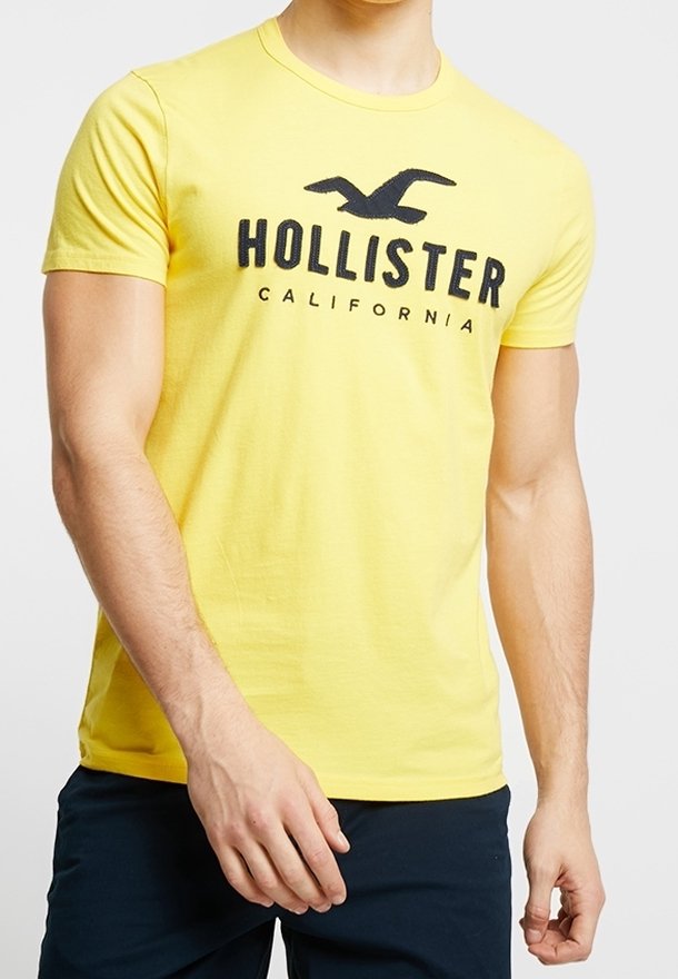 Gul bomullströja med korta ärmar, med en svart "HOLLISTER CALIFORNIA"-logotyp och en fågelgrafik på framsidan.