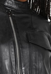 Veste en cuir noir avec un col montant, fermeture éclair à l'avant et poche à rabat. Texture lisse avec des détails de couture le long des coutures.