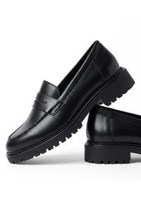 Par de mocasines negros de cuero con suelas gruesas y con dibujo, sobre fondo blanco, un zapato apoyado en el otro.