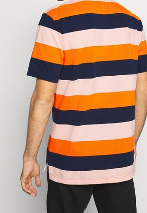 Man draagt een korte mouw polo met brede horizontale strepen in oranje, marineblauw en lichtroze, gezien van achteren.