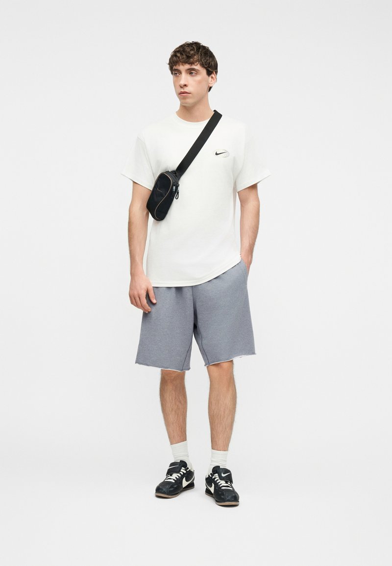 Νέος άνδρας που φοράει λευκό T-shirt Nike, γκρι σορτς, μαύρη τσάντα χιαστί, λευκές κάλτσες και μαύρα αθλητικά παπούτσια Nike, στέκεται μπροστά σε απλό φόντο.