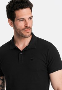 Černé polo tričko s krátkým rukávem, límečkovým designem, dvouknoflíkovou légou a vyšitým logem na levé části hrudi; strukturovaná tkanina.