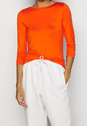 Femme portant un haut à manches longues orange vif et un pantalon blanc à cordon, avec une main dans la poche, sur fond uni.