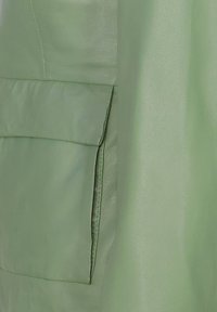 Groene leren kledingstuk met een gladde textuur, voorzien van een patchzak en een zijpaneel. Het ontwerp is eenvoudig en onopgesmukt.