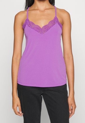 Vrouw die een paars, met kant afgewerkt hemdje met dunne bandjes draagt, gecombineerd met zwarte broek, staand tegen een effen achtergrond.