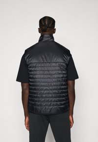Gepolsterte schwarze Weste mit hohem Kragen und horizontalen Nahtdetails, getragen über einem kurzärmeligen schwarzen Hemd. Glatte, glänzende Stofftextur.