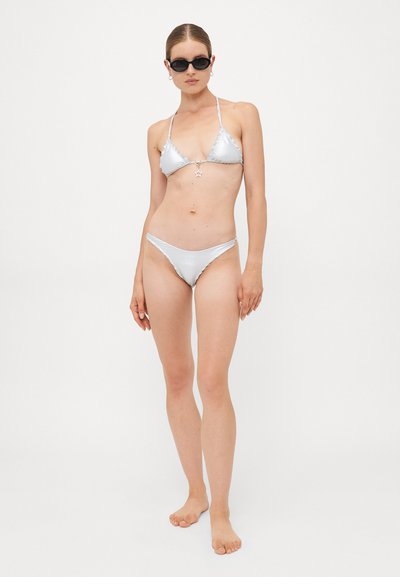 Woman wearing shiny silver bikini with star charm, black sunglasses, and small hoop earrings, standing barefoot against a white background.