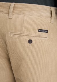Pantaloni in velluto a coste beige con righe verticali, dotati di una tasca posteriore, etichetta nera con scritte bianche e chiusura a un bottone.