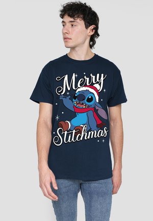 Junger Mann trägt ein dunkelblaues Shirt mit einem festlichen Stitch-Charakter, der einen Weihnachtsmann-Hut und einen Schal trägt, mit dem Text "Frohe Stitchmas."
