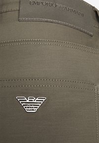 Khaki kalhoty s hladkou texturou, zdobené šedou koženou nášivkou v pase a stříbrným logem Emporio Armani na zadní straně.